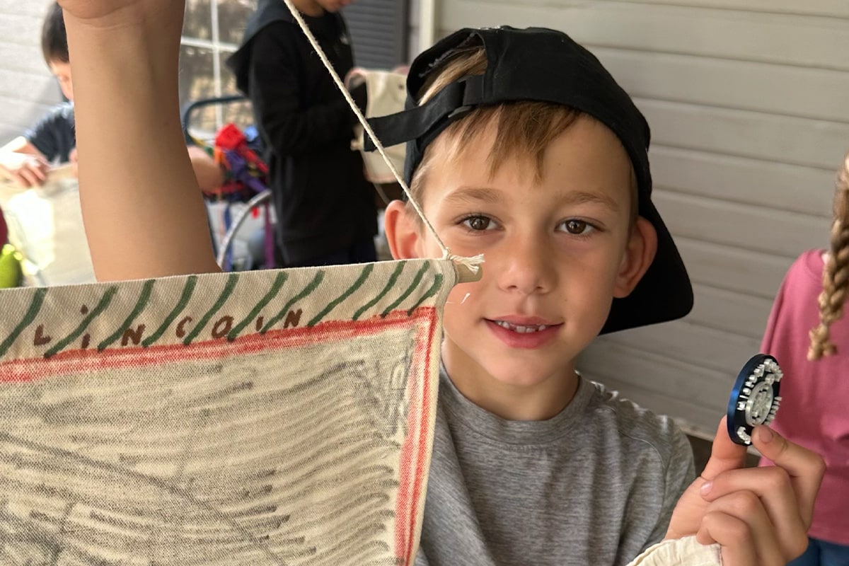 Young boy wearing black cap holds burlap sack and small object, smiling at camera during outdoor community event or festival activity.