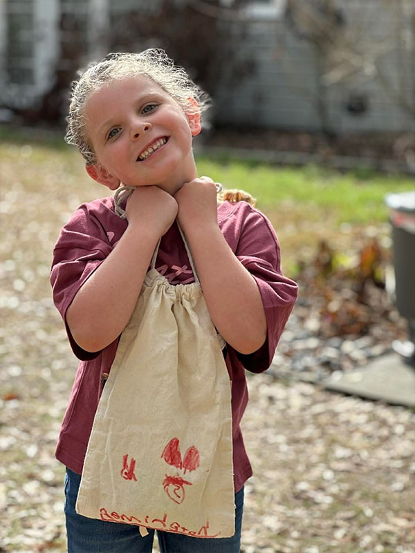 Happy young girl smiling outdoors wearing burgundy shirt and cream apron with red drawings, hands clasped under chin in sunny backyard