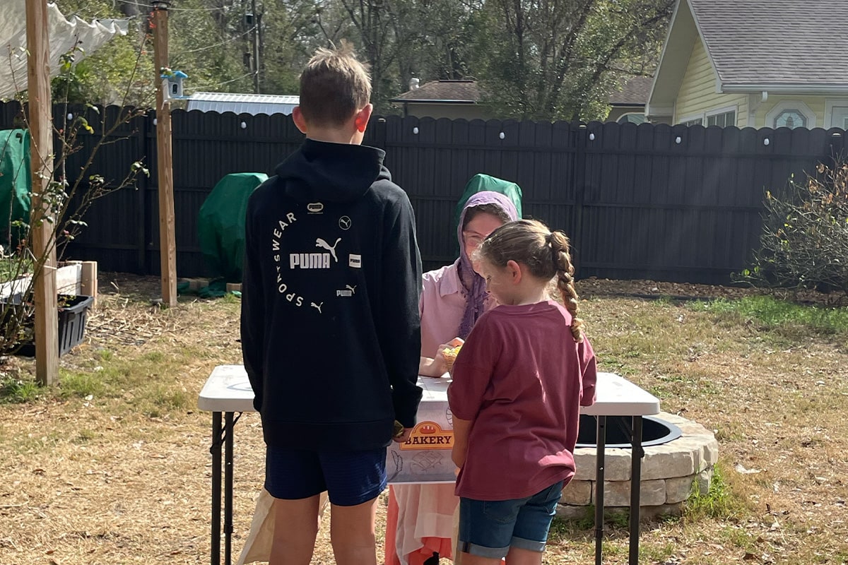 Three children gather around a white folding table in a backyard, with one wearing a black Puma hoodie and another in a red shirt with braided hair.